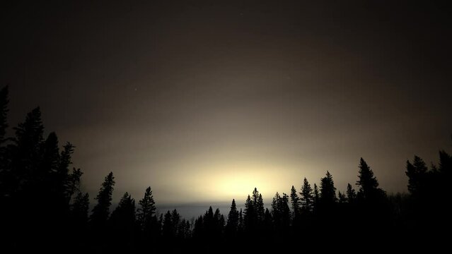 A night sky time lapse of white and gray clouds moving above a foreground of silhouettes of spruce and pine trees. A few stars can be seen through the clouds.
