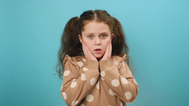 Omg. Portrait of preteen girl child opening mouth and touching face, looking at camera in shock, posing isolated over plain blue color background wall in studio. People emotion lifestyle concept