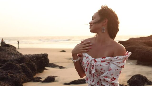Video close up smiling, sensual curly brunette female with shell bracelet, in white dress resting, relaxing at sea beach