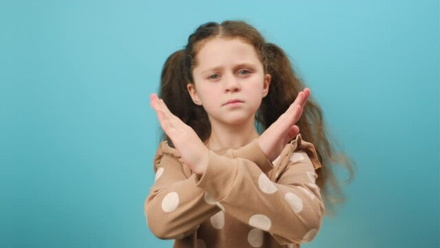 Portrait of serious preteen girl gesturing stop crossing hands, forbidding something, looking at camera, posing isolated over plain blue color background wall in studio. Rejection and no concept