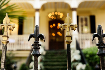 A cast wrought iron fence lined with black and gold fleur de lis post toppers with a New Orleans southern style home in the background