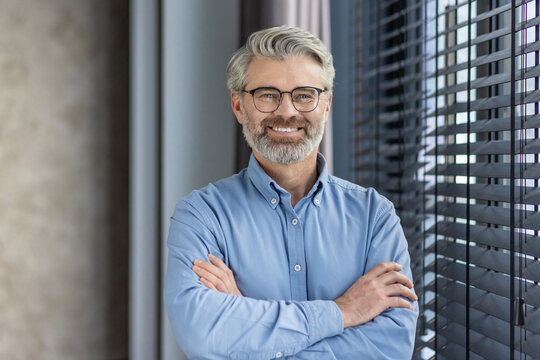 Portrait of mature successful businessman boss, senior gray-haired man inside office smiling looking at camera with crossed arms, experienced financier banker accountant in shirt.