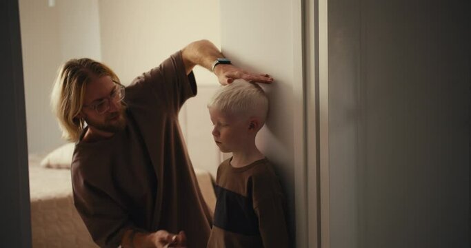 A little albino boy with white hair stands near the wall while his blond dad with a beard and glasses measures his height and then shows the boys height to him. Measure your childs growth