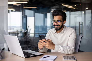 A young Indian man is sitting in an office at a desk and smilingly using a mobile phone