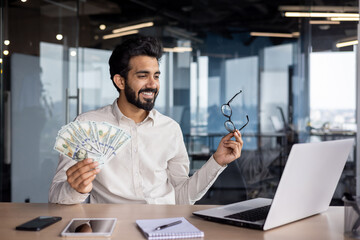 Happy young Muslim man sitting in office in front of laptop, talking on video call, showing and holding fan of money and glasses, bragging about achievement and success