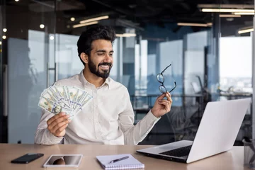 Fotobehang Happy young Muslim man sitting in office in front of laptop, talking on video call, showing and holding fan of money and glasses, bragging about achievement and success © Tetiana