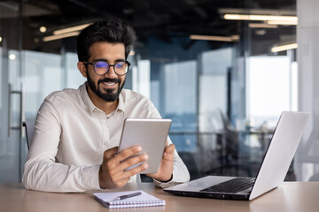 A smiling young Indian man in glasses is sitting in the office at a desk with a laptop and using a...