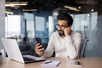 Fototapeta na wymiar Upset young Indian male businessman sitting at desk in office leaning tiredly on hand and looking frustrated at phone screen
