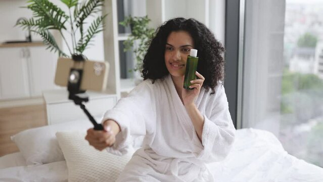 Beautiful young African woman social media influencer in bathrobe streaming and introducing beauty products to audience with phone holding skin moisturizing lotion.
