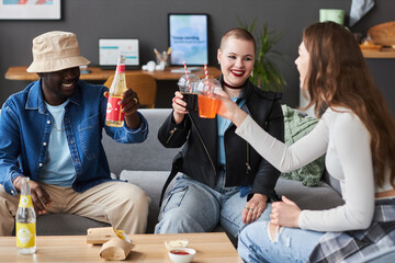Medium full shot of three cheerful diverse young people toasting with soda while sitting on couch...