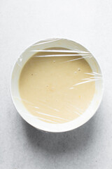 Vanilla madeleine batter in a white bowl covered with plastic wrap, a mixing bowl with thick...