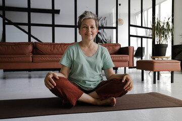 A middle aged lady rests after practicing yoga in the large hall of the house. A smiling woman sits on a mat on the floor in the lotus position in a happy mood after pleasant workout. Looks at the