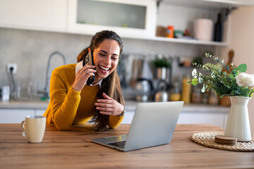 Smiling female entrepreneur discussing over mobile phone having entertaining conversation while sitting with laptop at kitchen counter. Young woman hearing good news over cell phone.