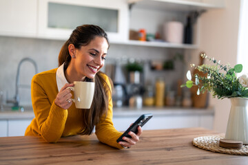 Focused businesswoman holding coffee cup and reading e-mails over smart phone sitting at kitchen...