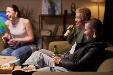 Side view at cheerful young girl with buzz cut sitting on couch drinking beer while socializing...