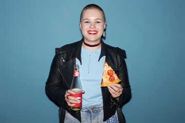 Medium shot of cheerful young girl with buzz cut holding slice of pepperoni pizza and bottle of...