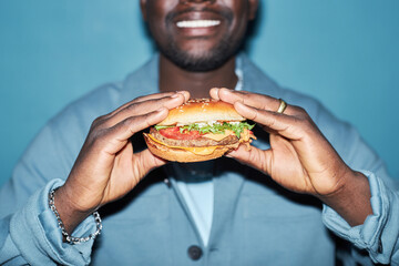 Close up shot of tasty meaty hamburger with vegetables in hands of Black man standing at light blue...