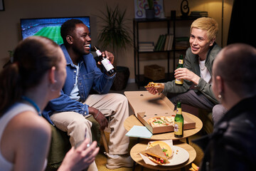 Medium full shot of diverse young adults sitting in living room laughing and talking while drinking...
