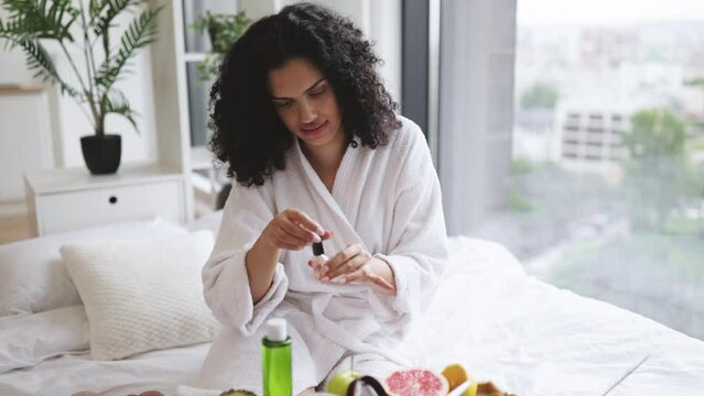 Portrait of female in white bathrobe making manicure while prepared ingredients for skin care in bright bedroom of apartment. Joyful housewife increasing life satisfaction by paints nails with polish.