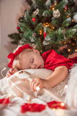Obraz na płótnie Canvas Cute little two year old baby girl in beautiful red dress sleeping under a Christmas tree, with decorations and lights around. Blonde hair, blue eyes. Presents, teddy bear. Taking a nap.