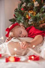 Obraz na płótnie Canvas Cute little two year old baby girl in beautiful red dress sleeping under a Christmas tree, with decorations and lights around. Blonde hair, blue eyes. Presents, teddy bear. Taking a nap.