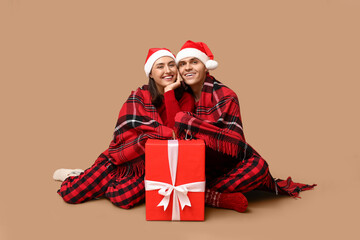 Happy young couple in Santa hats, with plaid and Christmas gift on beige background