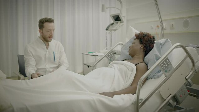 Young African Woman With Curly Hair Lying in Clinic Bed Talking to Physician