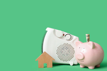 Electric heater with piggy bank, money and wooden house on green background. Concept of heating...