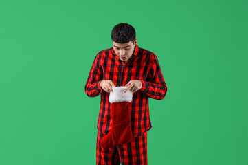 Young man in pajamas with Santa sock on green background