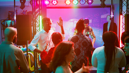 Diverse people partying with DJ on stage, young man mixing electronic sounds for crowd on dance floor. Friends dancing and having fun at underground disco party. Handheld shot.