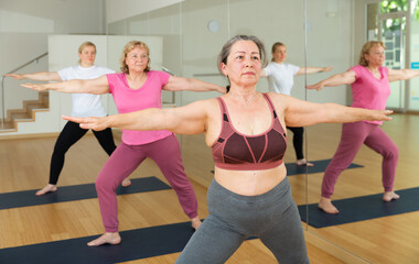 Three European ladies are doing warrior II pose beside a mirror