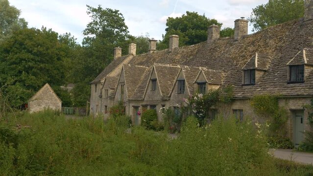 Romantic old stone cottages in iconic Bibury village on a cloudy summer day. Picturesque row houses peek out among the lush greenery in British countryside. Charming tourist destination worth visiting