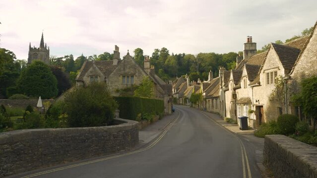 Entering a fairy tale country village with charming stone houses of honey colour. Picturesque terraced cottages placed along winding asphalt road below a green hill in delightfully pretty Castle Combe