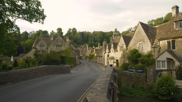 Crossing bridge towards idyllic countryside village with charming stone houses. Picturesque terraced cottages placed along a winding asphalt road below a green hill in delightfully pretty Castle Combe