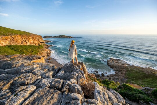 Young woman standing on a rock above the sea in the evening light, beach at The Gap, Robberg Peninsula hiking trail, Garden Route, Robberg Peninsula, Plettenberg Bay, Western Cape, South Africa