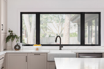 A kitchen detail with a black faucet on a white farmhouse sink in front of black framed windows, brown cabinets, and white marble countertops.