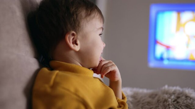 Adorable Caucasian toddler puts the finger into the mouth. Close up. Sweet kid watching TV at home.