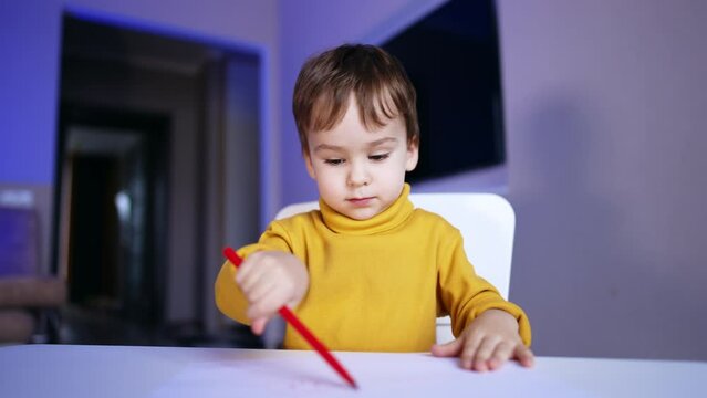 Focused baby boy drawing on the paper with a red pen. Lovely toddler learning to use felt pen at home.