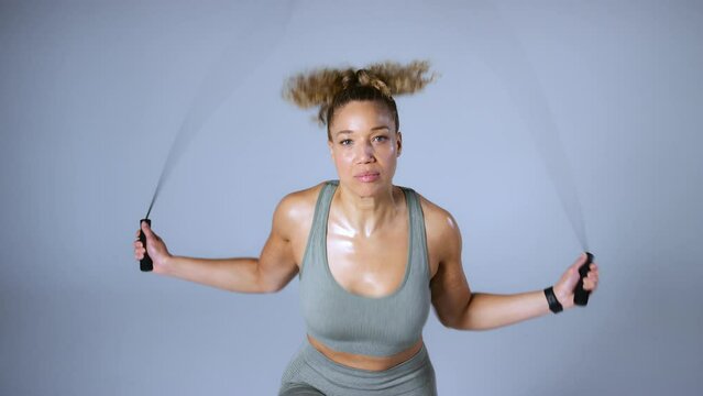 Studio shot looking down on smiling woman wearing gym fitness clothing exercising with skipping rope on grey background - shot in slow motion