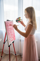 Young woman artist with palette and brush painting abstract pink picture on canvas near window. Art...