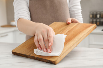 Woman wiping wooden cutting board with paper napkin at white table, closeup