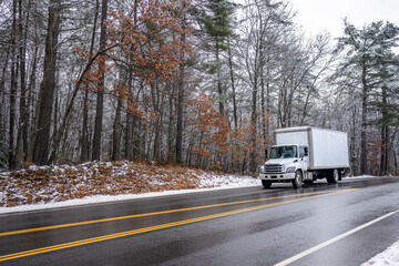 Fototapeta na wymiar Middle duty white day cab rig semi truck with box trailer for local freight driving on the winter slippery road with snow and ice and snowy forest on the side