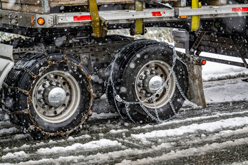 Properly and poorly secured chains for better grip on slippery road surfaces on wheels of the drive axles of semi truck carrying cargo on flat bed semi trailer in winter weather during snow storm - obrazy, fototapety, plakaty