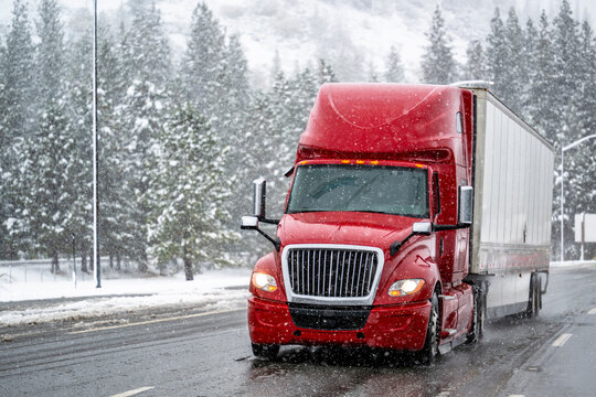 Tilted on the bend of the road red long hauler big rig semi truck with dry van semi trailer running on highway with forest and mountain on the side at the winter snowy storm time