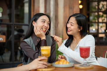 A lovely Asian girl is feeding food to her female friend, enjoying eating food at a cafe together.