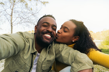 afro american couple taking a selfie kissing on the cheek. sitting in a park
