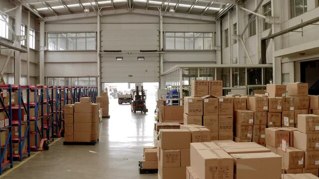 A forklift is parked between boxes at a warehouse of a Chinese factory. Hundreds of packaged products are organised on shelves at the storage facility Lots of sunlight is coming in through the windows