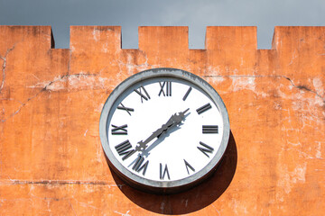 A round big clock with roman numbers in classic style that installed on the orange concrete wall. Vintage object photo.
