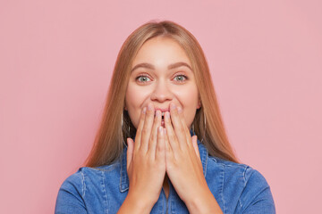 Golden-haired girl in blue denim dress holds hand at her face covering her mouth, good mood concept, copy space