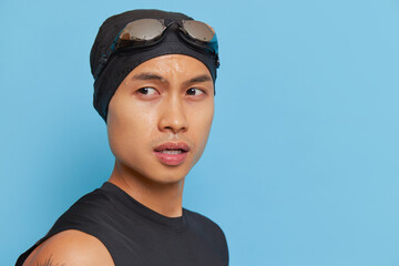 Portrait of sportive young man in swimming cap with goggles standing on blue background looking...